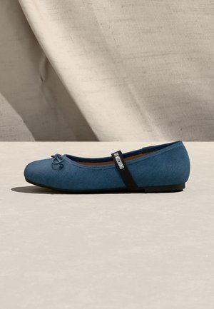Blue ballet flat with black elastic strap labeled "LOVE MOSCHINO" and small bow detail, set against a light beige textured background.
