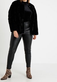 Eine Person trägt einen schwarzen Kunstfellmantel, schwarze Lederhosen, ein schwarzes Oberteil und Ankle Boots mit Leopardenmuster, während sie vor einem einfarbigen Hintergrund steht.