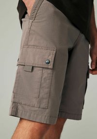 Närbild av en person som bär ljusbeiga cargoshorts med en stor klaffficka och tryckknapp i svart på vänster lår.
