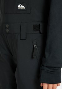 Sort snowboarddragt med tekstureret stof, dobbelt knaplukning, justerbar velcro-rem og en lynlåslomme på siden.