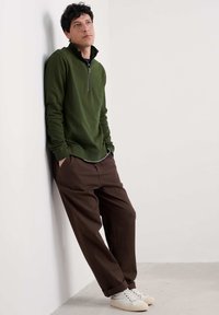 Hombre con sudadera verde de media cremallera y pantalones marrones, con las manos en los bolsillos, se apoya en una pared blanca, mirando hacia arriba. Zapatillas blancas visibles.