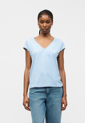 Kvinde med flettet hår iført lyseblå, tekstureret kortærmet V-hals top og jeans i mellemblå vask, stående mod hvid baggrund.