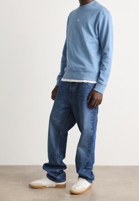 Sudadera azul claro con cuello redondo y logotipo blanco, combinada con vaqueros holgados azul oscuro y zapatillas blancas con suelas de goma.