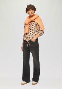 Lange zwarte jeans met een drawstring taille, een gestreepte blouse in oranje en zwarte geometrische vormen, en een oranje trui die over de schouders is gedrapeerd.