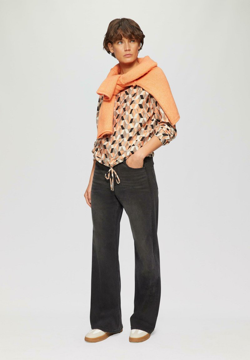 Lange schwarze Jeans mit einem Kordelzugbund, gemusterte Bluse in orange und schwarzen geometrischen Formen und ein orangefarbener Pullover, der über die Schultern gelegt ist.