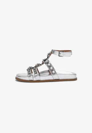 Sandal in argento metallico con più cinturini, abbellita con strass e una fibbia, caratterizzata da una suola piatta e una suola esterna color beige testurizzato.