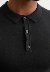 Polo noir en coton avec col et patte de boutonnage à trois boutons. Le tissu est lisse avec une texture côtelée autour du col.