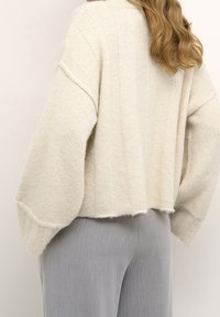 Jasno beżowy sweter wykonany na drutach o prążkowanej teksturze i opadających rękawach, z cropped długością i surowym wykończeniem. W zestawieniu ze szarymi spodniami.