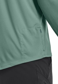 Camicia verde chiaro a maniche lunghe con una texture liscia, presenta una tasca laterale con cerniera e un orlo curvo. Abbinata a pantaloni neri.