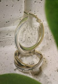 Sølv- og guldfarvede hoops øreringe med et snoet design, der har en poleret overflade og en hængselskonstruktion, præsenteret på en tekstureret overflade.