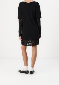 Zwarte tuniekjurk met korte mouwen en lange kantmouwen. Het kant heeft een patroon en de jurk heeft een rechte snit, tot halverwege de bovenbenen.