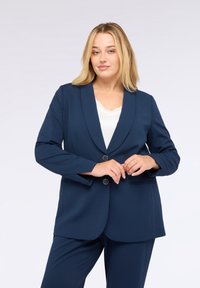 Kvinna med blont hår iklädd en marinblå kavaj och matchande byxor, poserande med händerna i fram. Ljus bakgrund.