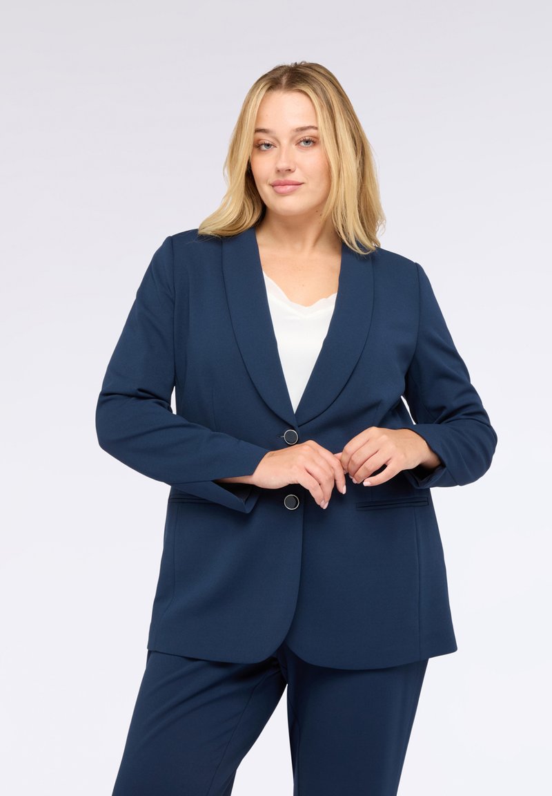 Kvinna med blont hår iklädd en marinblå kavaj och matchande byxor, poserande med händerna i fram. Ljus bakgrund.