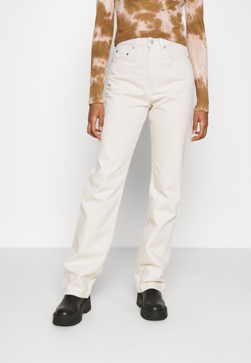 Jeans brancos de perna reta com uma cintura alta, feitos de denim. Combinados com um top de manga longa em tie-dye castanho e botas pretas de sola grossa.