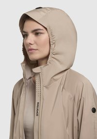 Jachetă cu glugă de culoare bej deschis, cu glugă strânsă, căptușeală din plasă și panou frontal perforat. Prezintă logo pe închizătoare și pe mânecă.