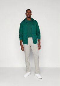 Groene zip-up hoodie met trekkoorden over een bijpassend T-shirt, gecombineerd met lichtgrijze joggingbroek en witte sneakers.