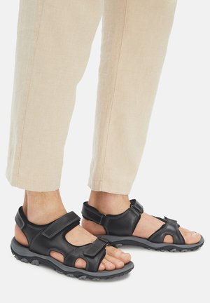 Schwarze Leder-Sandalen mit Klettverschluss, offenem Zehendesign und strukturierter schwarzer Gummisohle. Abgerundet durch kontrastierende weiße Steppnähte.