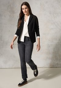 Blazer negro texturizado con dos botones, blusa blanca debajo, jeans gris ajustados y zapatillas negras con cordones blancos y suelas marrones.