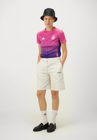 Maillot de sport dégradé rose et violet avec un motif triangulaire, associé à un short à rayures blanches. Chapeau seau noir et mocassins. Chaussettes blanches.