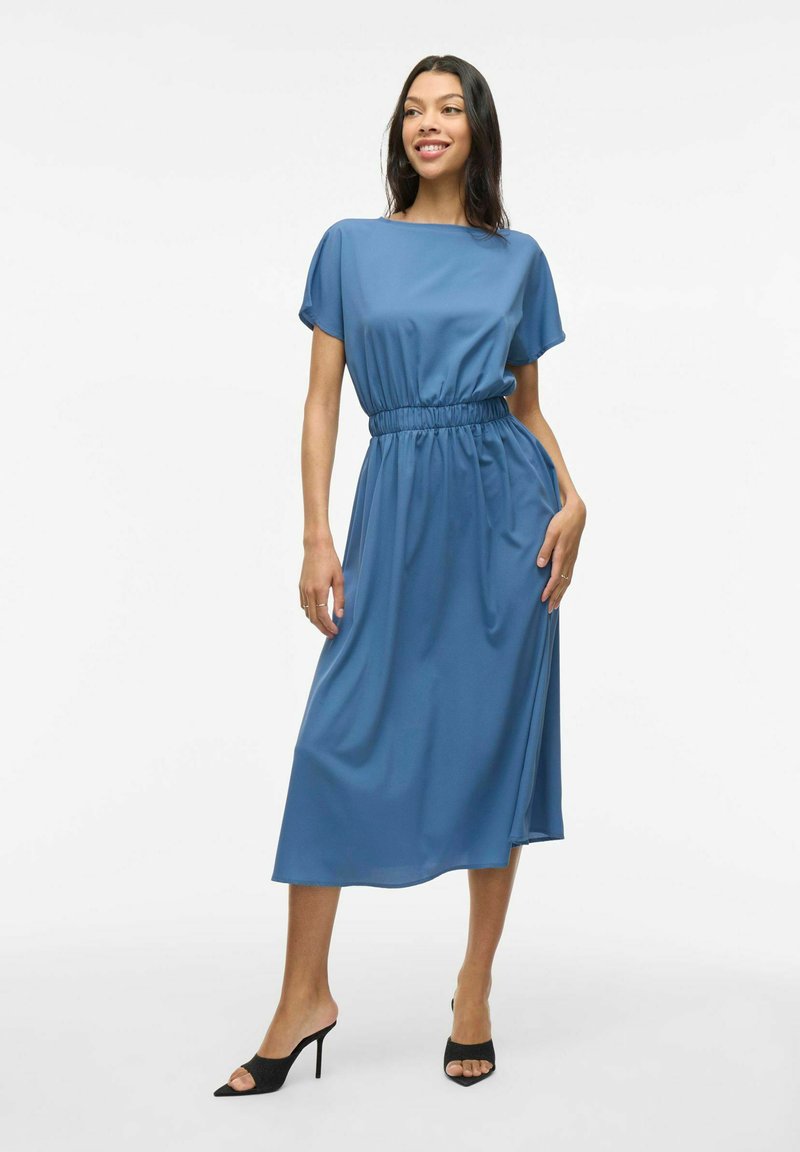 Rochie albastră, până la genunchi, dintr-un material neted și elegant, cu mâneci scurte și talie strânsă. Este purtată cu sandale negre cu vârf descoperit.
