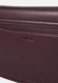Burgunderrød faux skinnveske med en glatt tekstur, med syddede detaljer og et preget "HVISK"-logo foran.