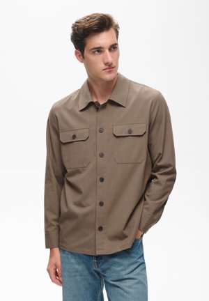 Jeune homme portant une chemise marron à boutons avec des poches poitrine et un jean bleu, regardant sur le côté avec une main dans la poche.
