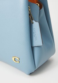 Light blue leather handbag with a textured finish, featuring a gold "C" logo and a small attached tag with brand name embossed.