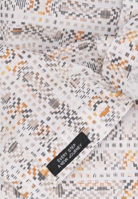 Witte stof met kleine oranje, beige en zwarte geometrische patronen en een zwart etiket met de tekst "IEDERE STAP EEN NIEUWE REIS."