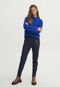 Kungsblå ribbad tröja med långa ärmar, kombinerad med marinblå slim-fit byxor med två guldfärgade dragkedjefickor. Bruna ankelboots med tjocka sulor.