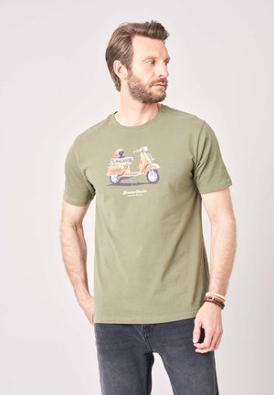 Man met baard die een olijfgroene T-shirt met oranje scooterprint en donkere spijkerbroek draagt, kijkt naar rechts.