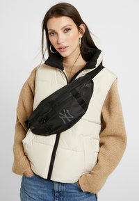 Sac banane noir en nylon avec logo, porté sur un gilet matelassé beige. Pull en maille texturé en dessous, jeans en denim bleu clair.