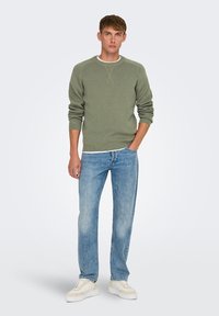 Maglione verde a maglia con texture a costine, camicia bianca a girocollo sotto, jeans blu a gamba dritta e sneakers beige. Sfondo semplice.