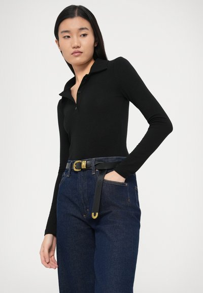 Naine, kes kannab musta pika varrukaga kraega särki, mis on sisse toppitud kõrge vöökohaga tumesinistesse teksadesse, millel on must vöö kuldse pandlaga.