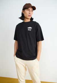 Black cotton t-shirt with a chest graphic featuring a car and the word "turbo." Worn with cream-colored pants, showcasing a relaxed fit.