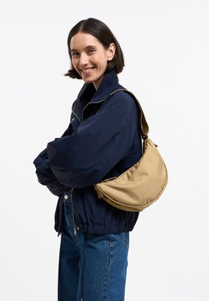 Donna sorridente, indossa una giacca blu navy e jeans blu, porta una borsa beige a tracolla sulla spalla sinistra su sfondo bianco.