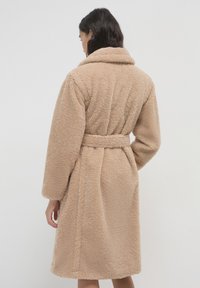 Donna con capelli scuri che indossa un cappotto teddy beige, lungo fino al ginocchio e con cintura, di spalle contro uno sfondo chiaro e semplice.