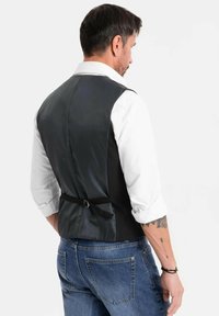 Uomo in camicia bianca con maniche arrotolate, gilet nero con cintura regolabile sul retro e jeans blu, di spalle contro uno sfondo semplice.