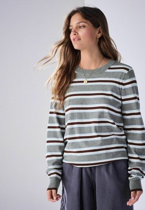 Ung kvinde med langt hår iført en grå stribet sweater og mørke bukser, kigger til siden mod en ensfarvet baggrund.