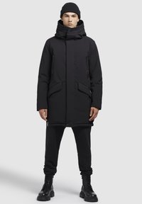 Sort isoleret parka med høj krave, to forlommer og lynlåse; parret med sorte sweatpants og snørede kampstøvler.
