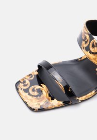 Μαύρο σανδάλι με τετράγωνη μύτη και χρυσό μπαρόκ μοτίβο στα λουριά και τη σόλα, με λεπτό μπροστινό λουράκι και λουράκι στον αστράγαλο.