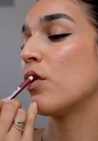 Läppennskrivare i burgunder appliceras på läpparna. Användaren har en neutral nagellack och lager av ringar i rosa, grönt och guld på sin hand.