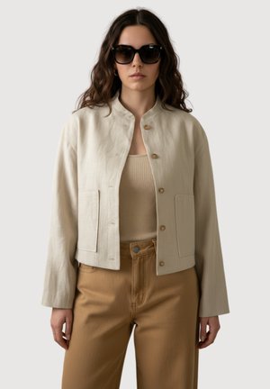Femme aux longs cheveux bouclés portant des lunettes de soleil noires, une veste courte beige, un débardeur côtelé et un pantalon taille haute beige, debout devant un fond uni.