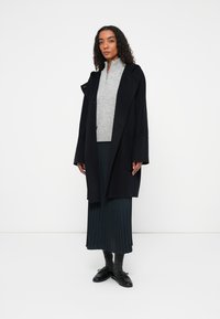 Černý oversized kabát s velkými klopami přes světle šedou zapínací svetr, doplněný plisovanou tmavozelenou sukní a černými botami.