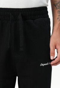 Black sweatpants with an elastic waistband, drawstring, side pockets, and white embroidered text "Originals" on the leg. Soft, textured fabric.