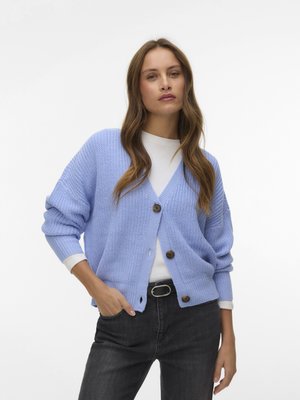 Femme portant un cardigan à gros tricot bleu clair sur un haut blanc et un jean noir avec une ceinture noire, debout avec une main dans la poche.