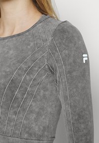 Graues sportliches Langarmoberteil, mit strukturiertem Stoff und erhabenen Nahtmustern, gerippten Bündchen und einem kleinen weißen Logo auf der Schulter.