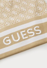 Bež kapa s uzorkom ima bijeli horizontalni pojas i izvezenu riječ "GUESS" u bijeloj boji.