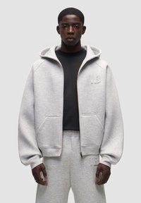 Felpa grigia con zip, dotata di ampie tasche, cappuccio regolabile e dettagli ricamati. Abbinata a pantaloni sportivi grigi coordinati. Tessuto morbido e texture.