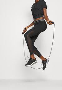 Conjunto atlético negro que incluye una camiseta corta y leggings capri con detalles de malla. Zapatillas negras con acentos verdes. Cuerda para saltar en la mano.