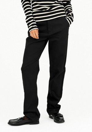 Personne portant un pantalon noir droit, des mocassins noirs et un haut à manches longues rayé horizontal noir et blanc avec une main dans la poche.